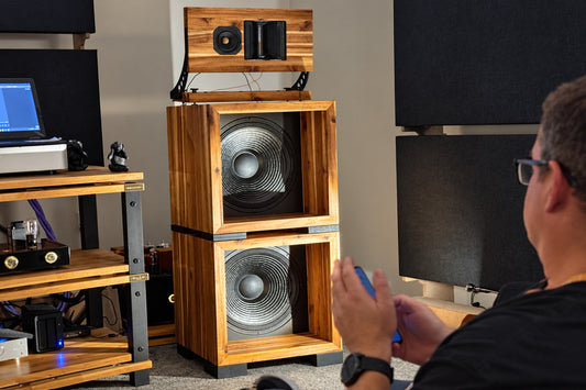 Visitor seated beside a wooden loudspeaker system and valve amplifier at an audio show.